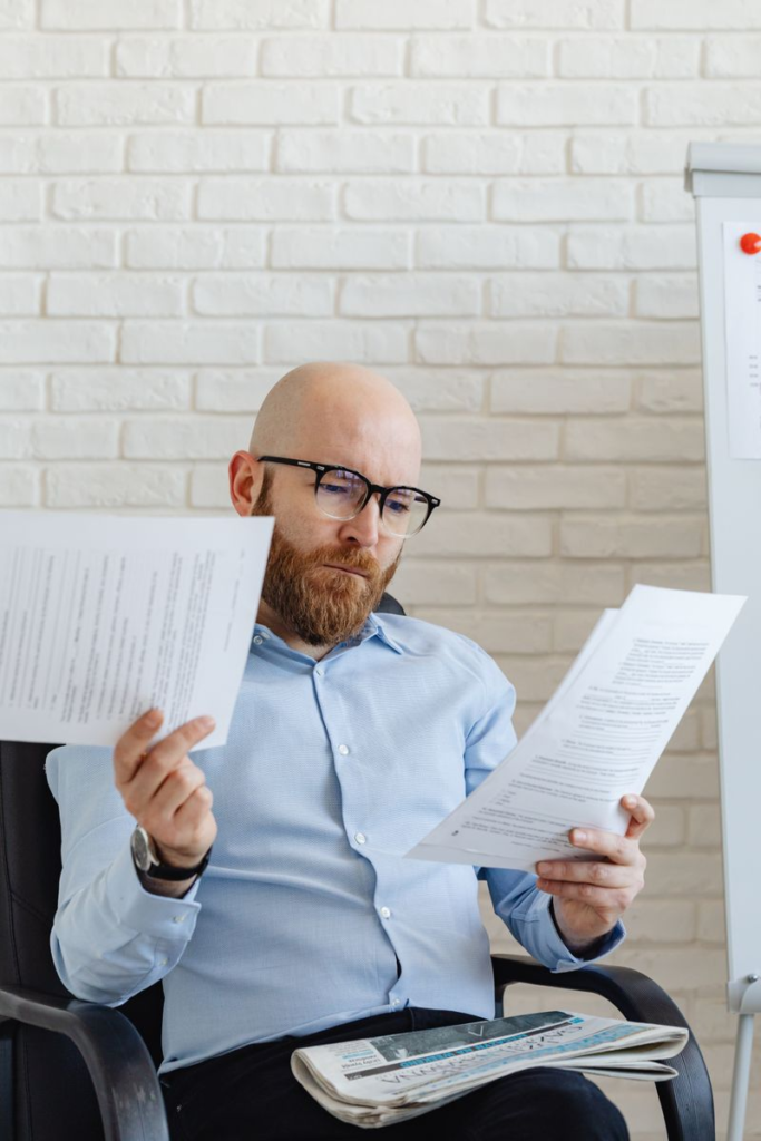 A serious-looking bald man with glasses reviews papers in an office against a brick wall.